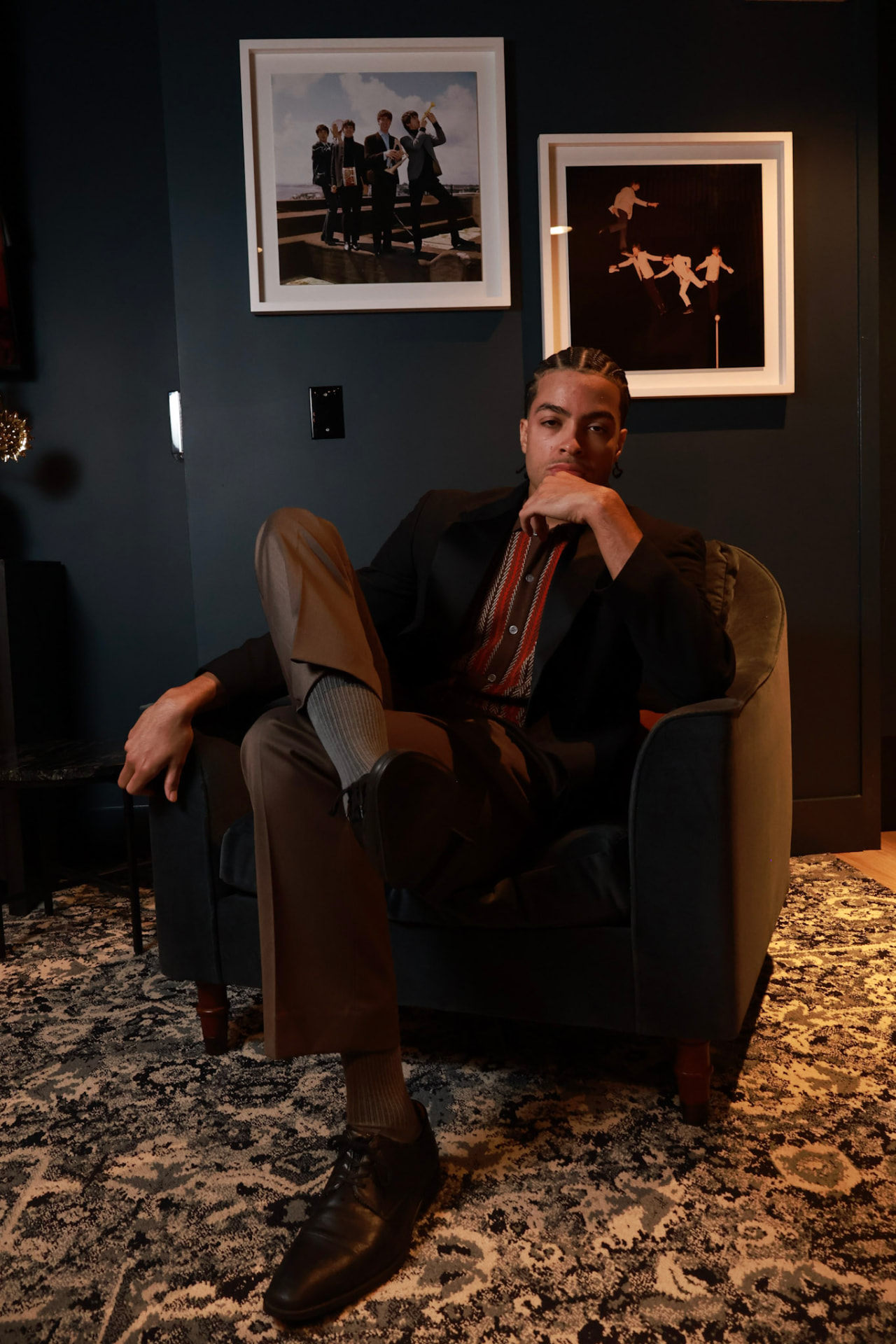Portrait of a man with braided hair in a dark blazer and patterned shirt, seated in an armchair