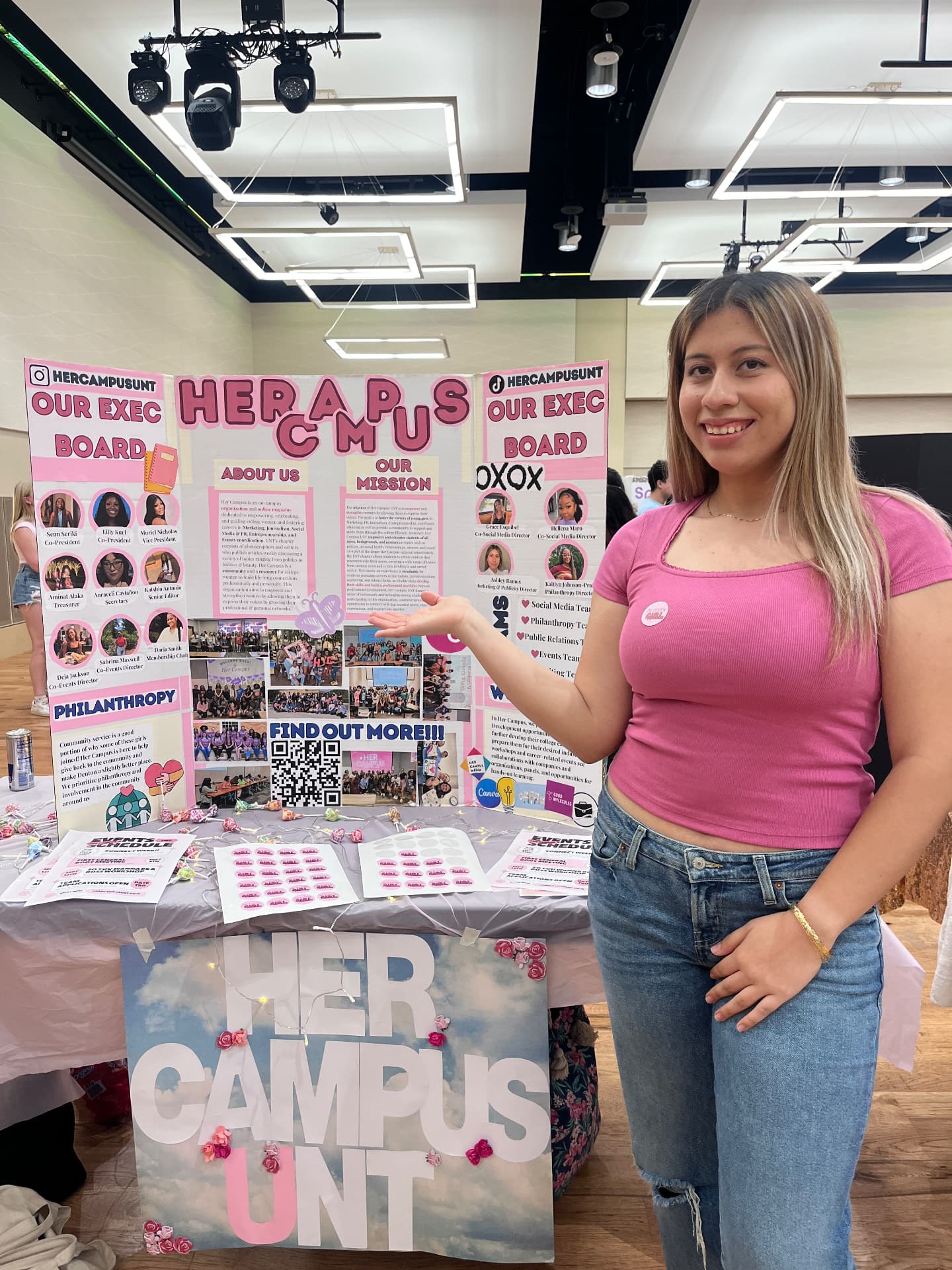 A woman in a pink t-shirt gesturing toward a pink Her Campus UNT tri-fold display board at a campus recruitment fair, with flyers and a table sign in front